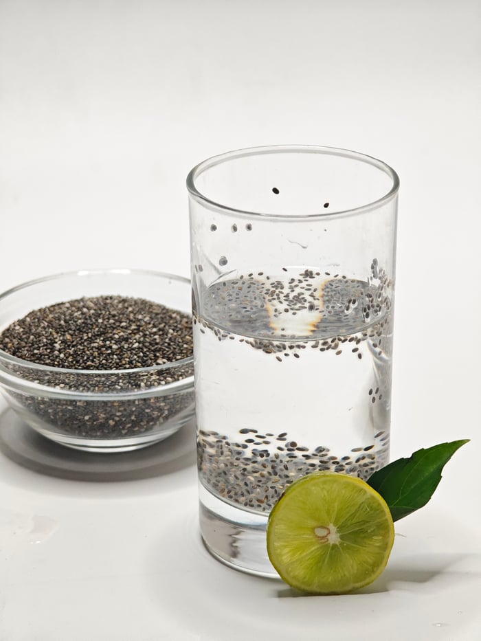 Glass of chia seed water with a lime slice and bowl of seeds on white surface.